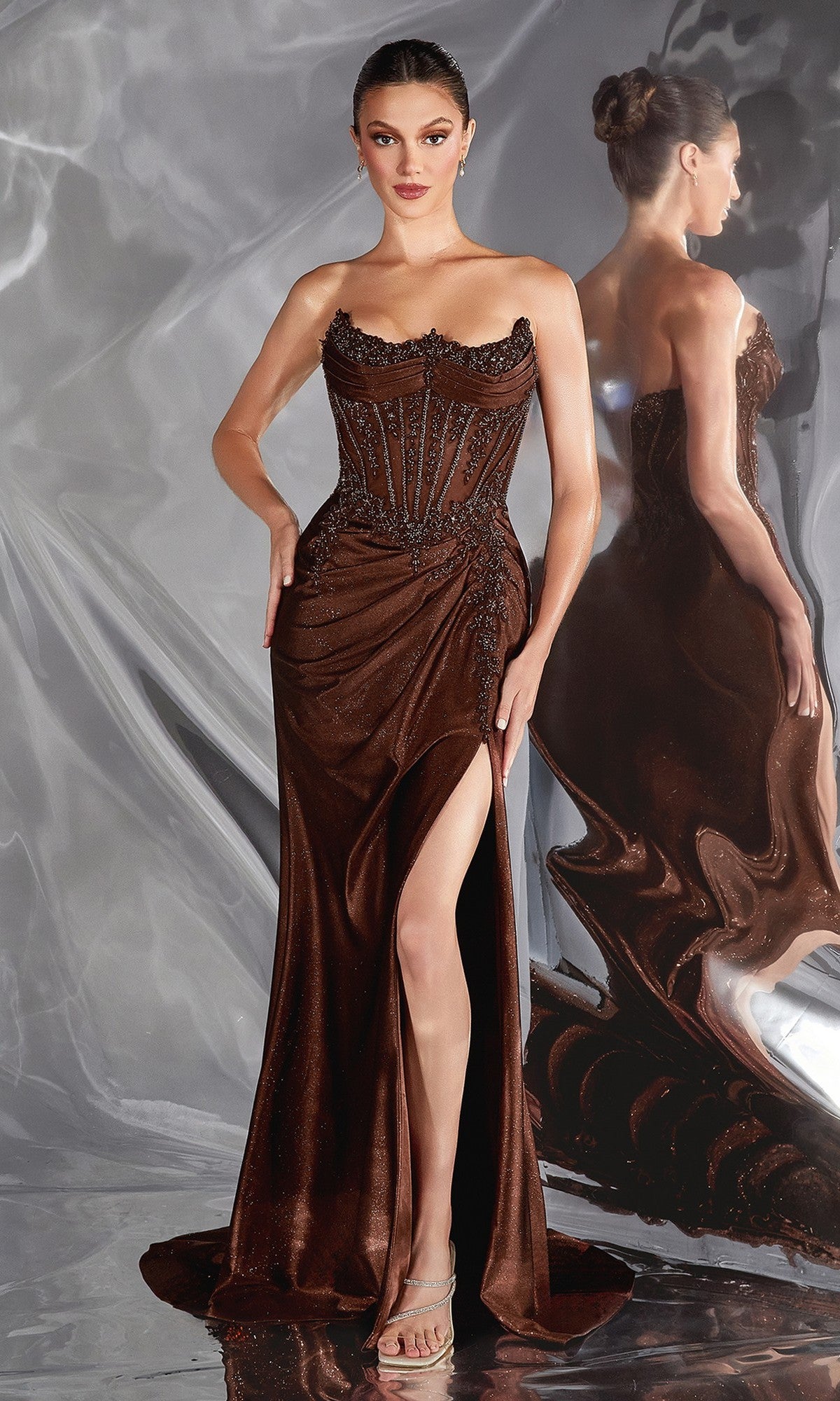 A woman in the Sheer-Corset Long Prom Dress: Ladivine CC463, featuring a strapless bodice with a reverse-sweetheart neckline and thigh-high slit, stands before a reflective backdrop, her elegant reflection beside her.